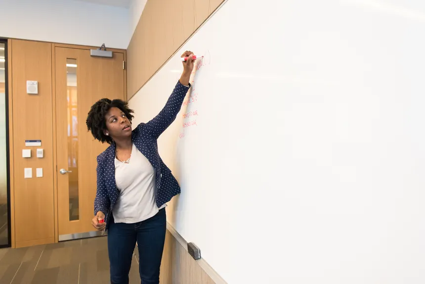 Female teacher writing on a whiteboard in a classroom, explaining a lesson to students.