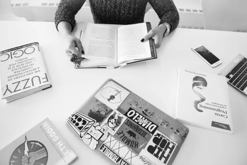 Person reading amidst books and technology on a table in a monochrome setting.