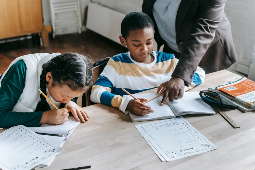 High angle of crop unrecognizable black female teacher explaining task to focused little schoolboy sitting at desk near attentive Asian classmate writing in not