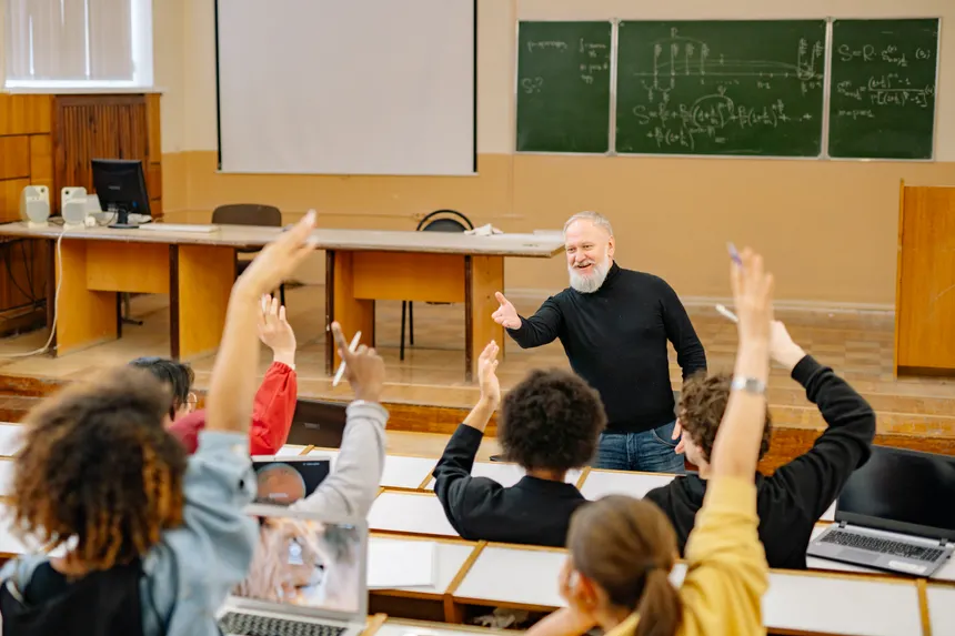 Professor engaging with students in a lively university classroom discussion as students raise hands.